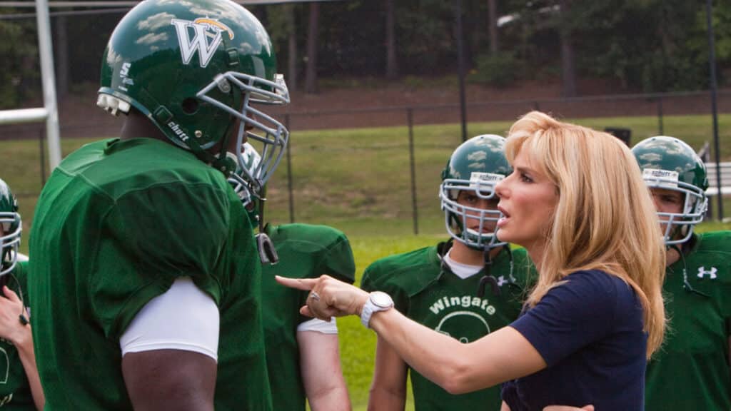 Screenshot from The Blind Side showing Leigh Anne Tuohy standing beside Michael Oher on a football field, with Oher in uniform and Tuohy dressed casually, emphasizing their mentor-guardian dynamic.