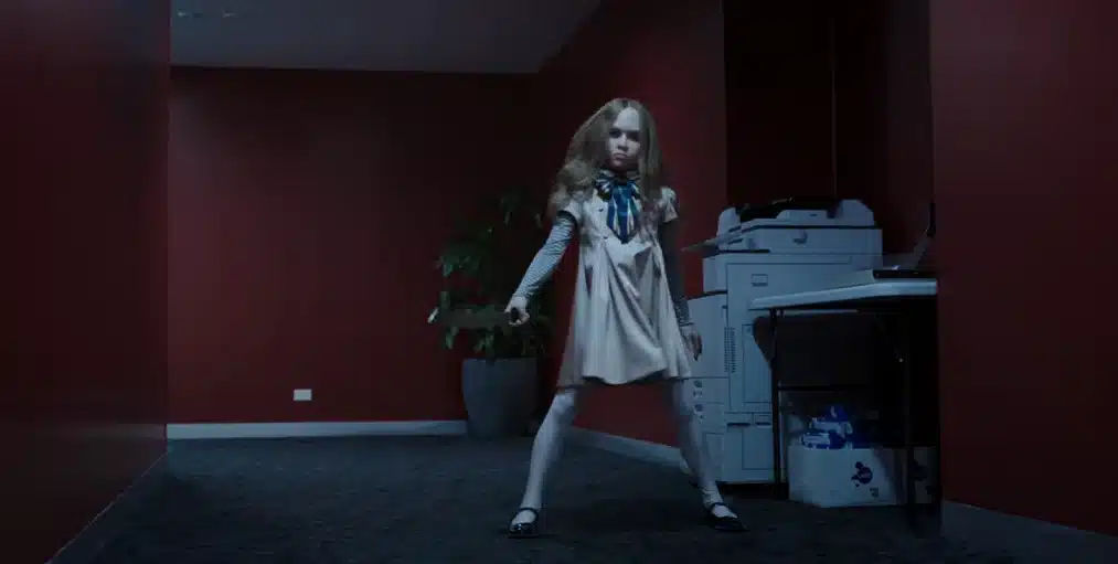 M3GAN, the lifelike AI doll, standing in a hallway and staring forward.